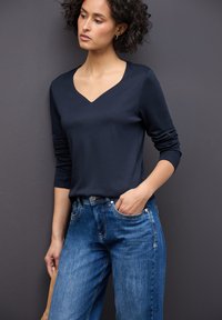 Top blu navy a V con maniche lunghe, caratterizzato da una texture liscia. Abbinato a jeans a gamba larga di un blu chiaro, che mostrano una finitura denim classica.