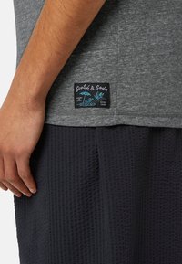 Tricou gri cu un material texturat, având un patch cu logo-ul Scotch & Soda în culorile negru și albastru pe partea de jos, asortat cu pantaloni scurți texturați de culoare închisă.