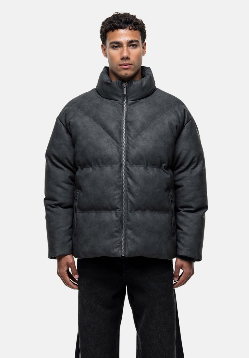 Mann in einer dunkelgrauen gepolsterten Jacke mit hohem Kragen und seitlichen Reißverschlusstaschen, kombiniert mit schwarzen Hosen, gegen einen schlichten Hintergrund stehend.