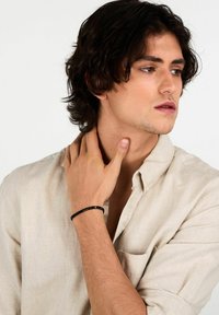 Bracciale nero con perline su un polso, caratterizzato da perline lisce e rotonde. Il modello indossa una camicia beige chiaro con bottoni e si tocca il collo.