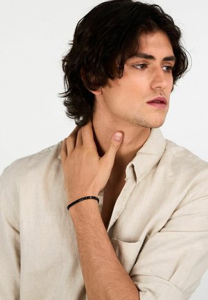 Bracciale nero con perline su un polso, caratterizzato da perline lisce e rotonde. Il modello indossa una camicia beige chiaro con bottoni e si tocca il collo.