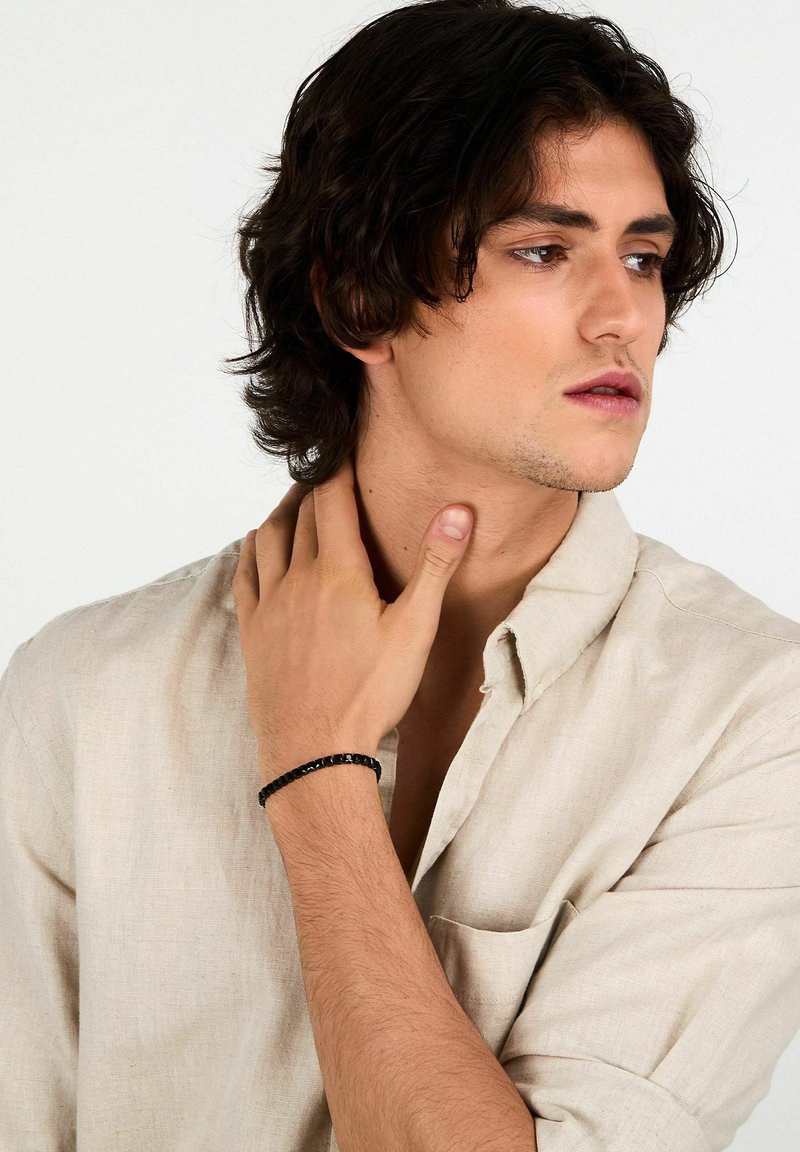 Bracciale nero con perline su un polso, caratterizzato da perline lisce e rotonde. Il modello indossa una camicia beige chiaro con bottoni e si tocca il collo.