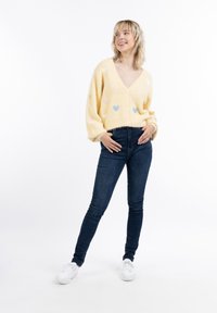Cardigan giallo soffice con scollo a V profondo, decorato con motivi a forma di cuore nei colori pastello. Indossato con jeans aderenti scuri e sneakers bianche.