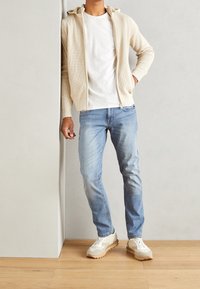 Beige zip-up hoodie with textured fabric, white t-shirt underneath, light blue jeans, and light-colored sneakers with a thick sole.