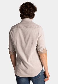Lichtbeige overhemd met lange mouwen en opgerolde manchetten, gemaakt van een eenvoudige, gestructureerde stof, met een klassieke kraag en een gebogen zoom, gedragen over blauwe jeans.