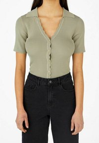Geribbelde blouse met korte mouwen in olijfgroen, met een gekartelde kraag en knopen aan de voorkant. Gecombineerd met hoge zwarte spijkerbroeken.
