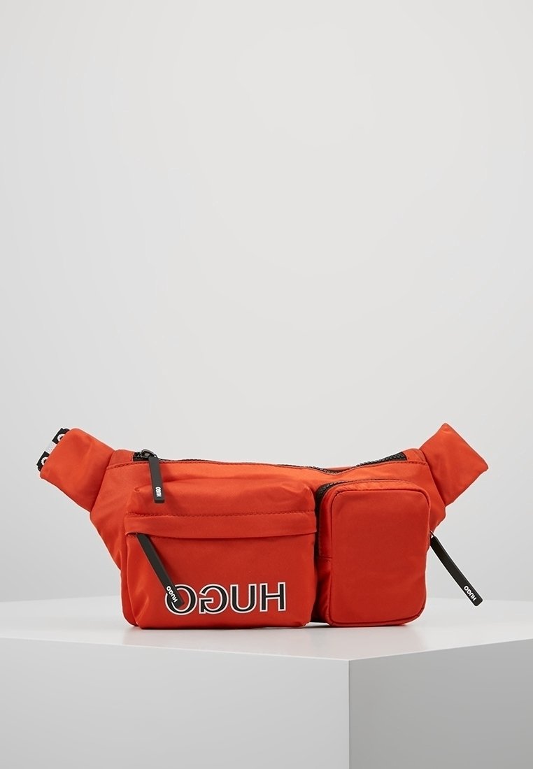 Banane orange vif avec plusieurs compartiments zippés et logo "HUGO" sur la poche avant, présentée sur une surface blanche.