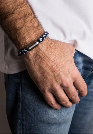 Mannenhand rust op de heup, gekleed in een blauwe spijkerbroek en een wit shirt, met een blauw-wit gevlochten armband met een zilveren sluiting om de pols.