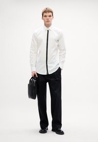 Witte lange mouw shirt met een zwart voorpaneel, gedragen met losse zwarte broek en gepolijste zwarte schoenen. Draagt een zwarte tas.