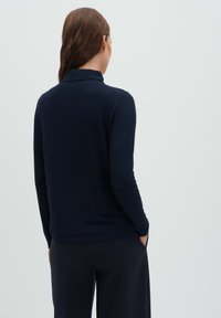 Donna con capelli lunghi castani che indossa una maglia a maniche lunghe blu navy e pantaloni blu navy, in piedi con le mani nelle tasche, di spalle su uno sfondo semplice.