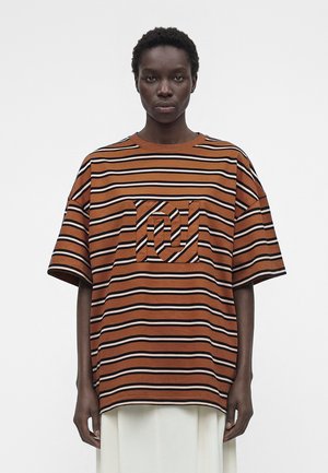 Persoană care poartă un tricou larg în dungi portocaliu rugină și negru cu un buzunar pătrat în piept și pantaloni albi cu picioare largi, stând în fața unui fundal alb.