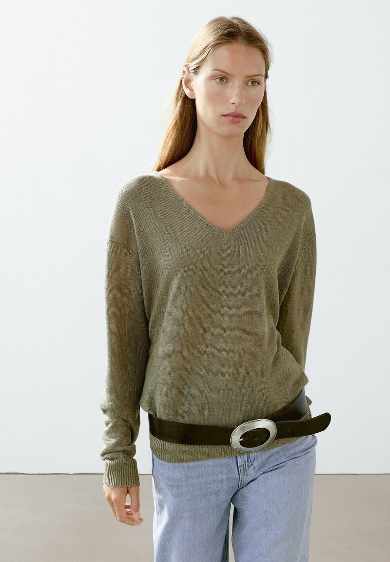 Olivengrüner V-Ausschnitt-Pullover mit langen Ärmeln, gestrickte Textur. Trägt einen schwarzen Gürtel mit einer großen silbernen Schnalle. Hellblaue Jeans.