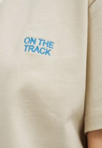 Μπεζ ύφασμα με μπλε κεντημένο κείμενο "ON THE TRACK" κοντά στην περιοχή του άνω στήθους ενός ενδύματος με κοντά μανίκια.