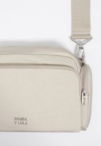 Beżowa prostokątna torebka na ramię o gładkiej fakturze, z zamykanymi na zamek przegrodami i szerokim regulowanym paskiem. Z metalowymi srebrnymi dodatkami.