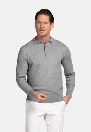 Man draagt een lichtgrijze polo met lange mouwen en witte broek, staand met één hand in de zak tegen een effen achtergrond.