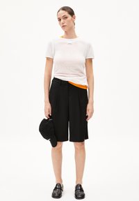 White ribbed short-sleeve shirt layered over an orange accent, paired with black pleated shorts. Model holds a black cap, wearing black loafers.