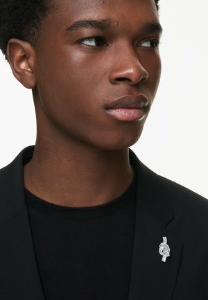 Black blazer over a black t-shirt, featuring a silver brooch with sparkling accents, positioned on the left side of the blazer.