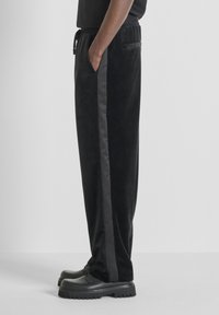 Zwarte velours joggingbroek met satijnen strepen aan de zijkant, trekkoord in de taille en zakken. Gecombineerd met stevige zwarte schoenen. Over het geheel een gladde textuur.