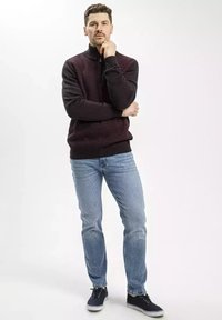 Ciemno bordowy i czarny sweter z dzianiny o wysokim kołnierzu, zestawiony z jasnoniebieskimi dżinsami i granatowymi sneakersami, stojący na jednolitym tle.