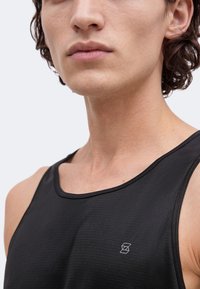 Jonge man met krullend haar, die een zwart mouwloos sportshirt draagt met een klein wit logo op de borst tegen een witte achtergrond.