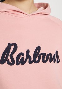 Różowa bluza z kapturem, z ciemnogranatowym haftowanym napisem "Barbour" na piersi, wykonanym ozdobną czcionką.