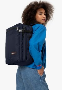 Mochila Eastpak de color navy con dobles cremalleras, correas ajustables y bolsillo frontal, hecha de tela duradera. El modelo posa de manera casual vistiendo una chaqueta azul.