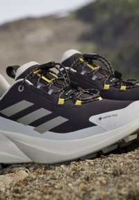 Chaussures de trail Gore-Tex noires et blanches avec accents jaunes et système de laçage rapide sur un terrain extérieur rocailleux.