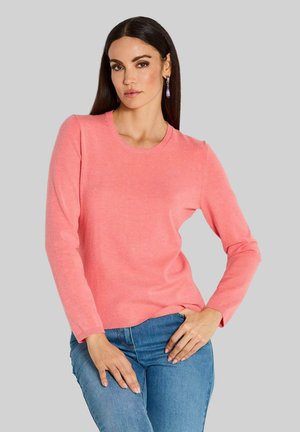 Rosa langärmliger Pullover mit rundem Halsausschnitt. Glatte, weiche Textur und gerippte Bündchen. Kombiniert mit enganliegenden blauen Jeans.