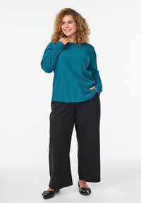 Lächelnde Frau mit lockigem Haar, die ein türkisfarbenes langärmliges Oberteil, schwarze weite Hosen und schwarze nietenbesetzte Ballerinas trägt und mit einer Hand in der Tasche steht.