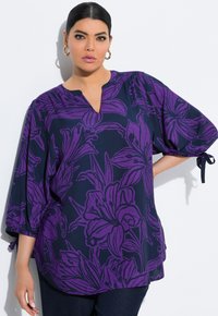 Navy floral bluse med store lilla blomstermønstre, en V-halsudskæring, og elastiske puffærmer med bindedetaljer ved manchetterne.