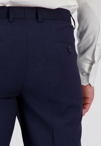 Man in een donkerblauwe op maat gemaakte broek met een knoopzak aan de achterkant en een witte overhemden met knoopmanchetten.