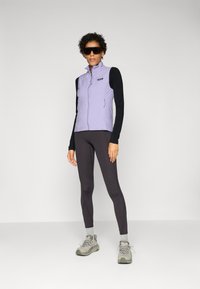 Vesta izolată de un violet deschis, cu fermoar frontal, asortată cu un top negru cu mânecă lungă și leggings închise la culoare. Purtați cu pantofi atletici gri.