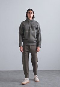 En ung mann med langt hår står i grå treningsdrakt og hvite sneakers mot en enkel lys grå bakgrunn og betonggulv.
