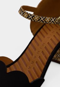 Sandale noire avec un bord festonné, bride de cheville à motifs en beige, marron et noir. Intérieur avec une semelle texturée beige. Accent en métal doré.