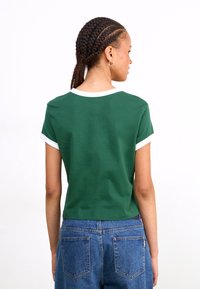 Green short-sleeve t-shirt with white trim at the neckline and sleeves. Fabric appears soft with a standard fit. Worn with high-waisted blue jeans.