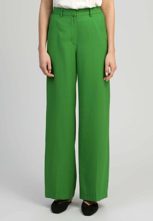 Femme portant un pantalon vert taille haute à jambes larges avec passants de ceinture, fermeture éclair à l'avant, et des ballerines noires pointues sur un fond uni.