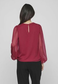 Donna che indossa una blusa bordeaux a maniche lunghe con maniche trasparenti e un piccolo foro con bottone sul retro, abbinata a pantaloni neri.