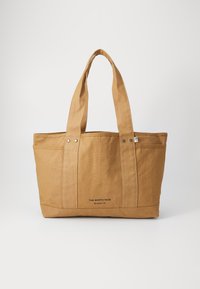 Tan canvas tote bag with dual handles, two rounded top seams, and a logo printed near the base. Features snap fasteners on the exterior.