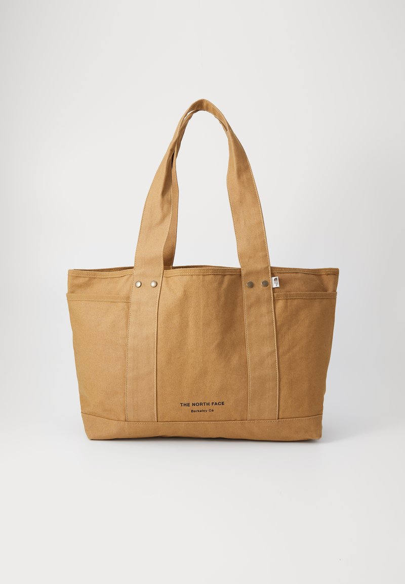 Tan canvas tote bag with dual handles, two rounded top seams, and a logo printed near the base. Features snap fasteners on the exterior.