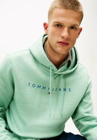 Mintgrøn hættetrøje med "TOMMY JEANS" i blå skrift og en lille logoaccent. Fremstillet af blødt stof med snøre i hætten.