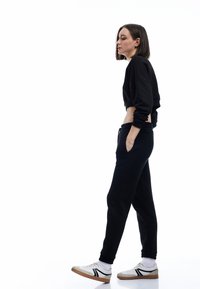 Młoda kobieta w czarnej, krótkej bluzy i dresach, w białych sneakersach, stojąca w profilu z rękami w kieszeniach na białym tle.