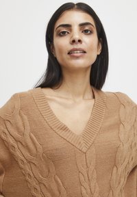 Tjockstickad tröja i varm beige, med V-ringning och flätade mönster på framsidan. Mjuk textur med ribbat detalj vid halsringningen.
