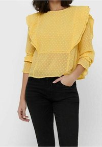 Gelbe Bluse mit gepunktetem Muster, Rüschensleeves, transparenter Stoff und lässiger Passform. Kombiniert mit schwarzen eng anliegenden Hosen für den Kontrast.