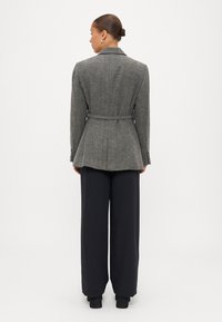 Blazer em padrão espinha de peixe cinza com um corte ajustado, cintura cingida e ombros estruturados, combinado com calças pretas de perna larga e botas.