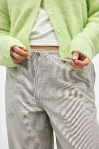 Zelený chlupatý cardigan s knoflíky přes bílou košili, doplněný šedými a bílými pruhovanými kalhotami na šňůrku. Viditelné detaily texturované látky.
