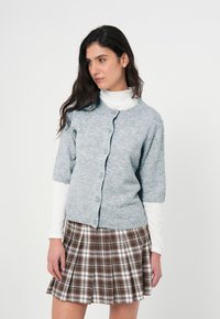 Ανοιχτό γκρι πλεκτή ζακέτα με κοντά μανίκια, κουμπιά στο μπροστινό μέρος και ριμπ μανσέτες, φορεμένη πάνω από λευκή μπλούζα με μακριά μανίκια και καρό καφέ φούστα.