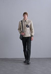 Μπεζ ζακέτα από μείγμα μαλλιού με μαύρα κουμπιά, που φέρει το λογότυπο "EA", με μαύρες ρίγες στις μανσέτες, φορεμένη πάνω από σκούρα παντελόνια και ένα κασκόλ καφέ.