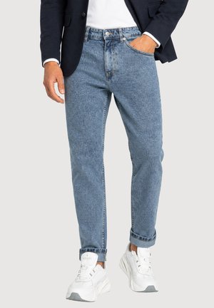 Uomo con jeans azzurri chiari risvoltati, sneakers bianchi, camicia bianca e blazer scuro con una mano in tasca.