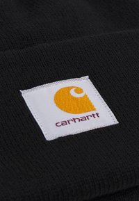 Melna trikotāžas cepure ar pielipinātu kvadrātveida baltu zīmi, kurā ir dzeltenais un melnais Carhartt logo. Audumam ir teksturēta virsma.