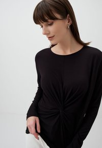 Zwarte lange mouw top met een gedraaid ontwerp bij de taille, ronde halslijn en een gladde textuur. Model poseert met een neutrale achtergrond.